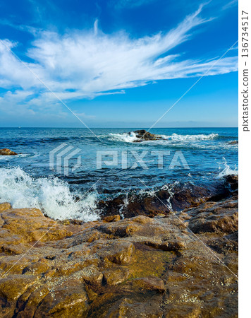 Waves crash on rocks near the shore under a blue sky with white clouds in the early afternoon Waves crash on rocks near the shore under a blue sky with white clouds in the early afternoon 136734517