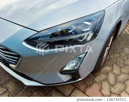 Close view of a silver car parked on a brick surface showing details of the front headlight and design elements during a rainy day Close view of a silver car parked on a brick surface showing details of the front headlight and design elements during a rainy day 136734551