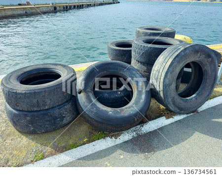 Old tires are stacked beside the water at a port during a sunny day in the afternoon, showing the effects of weathering and time on used rubber Old tires are stacked beside the water at a port during a sunny day in the afternoon, showing the effects of weathering and time on used rubber 136734561