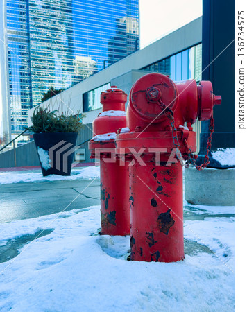 Two red fire hydrants stand in snow beside a modern building in a city downtown area during winter with tall glass structures in the background reflecting sunlight Two red fire hydrants stand in snow beside a modern building in a city downtown area during winter with tall glass structures in the background reflecting sunlight 136734575