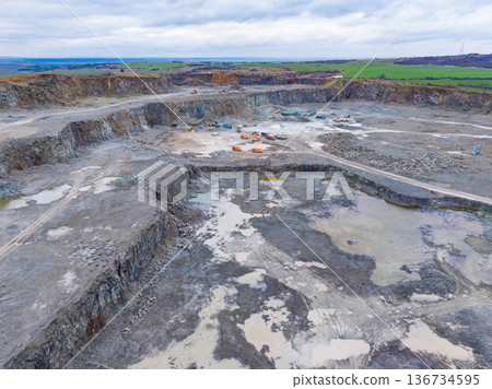 Construction site shows large quarry with machinery and vehicles moving dirt near small hills and cloudy sky in the background during daytime Construction site shows large quarry with machinery and vehicles moving dirt near small hills and cloudy sky in the background during daytime 136734595