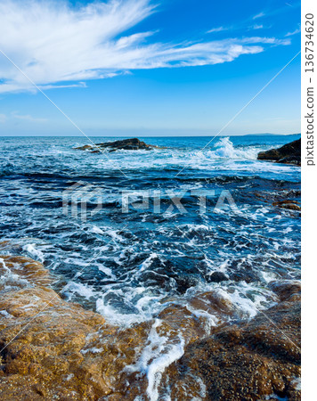 Waves crash on rocks along the coast under a blue sky with clouds in the afternoon 136734620
