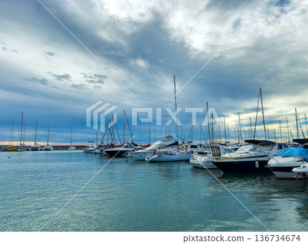 Boats are docked in a marina with cloudy skies above and calm waters during the late afternoon near the coast 136734674