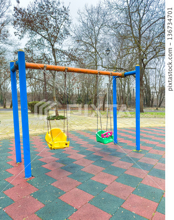 Swings are placed at a playground surrounded by trees on a cloudy day in the afternoon in a park setting Swings are placed at a playground surrounded by trees on a cloudy day in the afternoon in a park setting 136734701