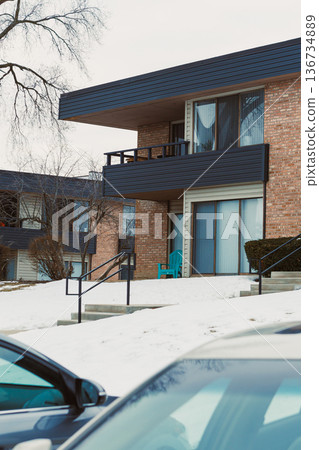 Winter scene showing an apartment building with a single chair on the balcony and cars parked in front 136734889