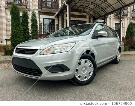 Grey sedan parked outside a large building with green trees near the entrance on a cloudy day Grey sedan parked outside a large building with green trees near the entrance on a cloudy day 136734900