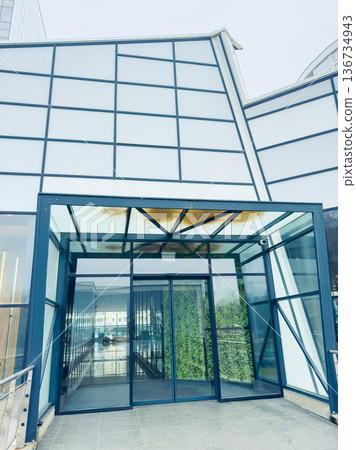 Modern building entrance with glass walls and greenery, showcasing a stylish design in an urban setting during daylight Modern building entrance with glass walls and greenery, showcasing a stylish design in an urban setting during daylight 136734943