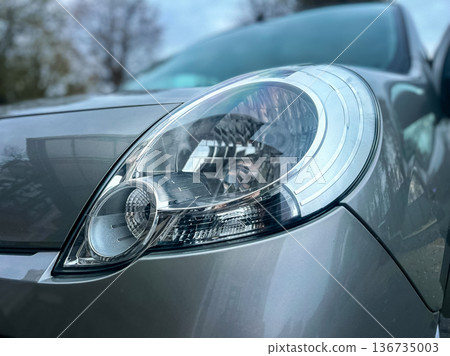 Close view of a car headlight in a parking area during the day with trees and a building in the background showing reflections on the surface Close view of a car headlight in a parking area during the day with trees and a building in the background showing reflections on the surface 136735003