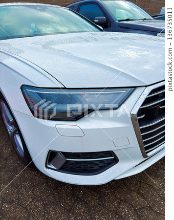 White car parked in a lot with rain droplets on the surface, showing part of the vehicle and nearby cars during a cloudy day 136735011