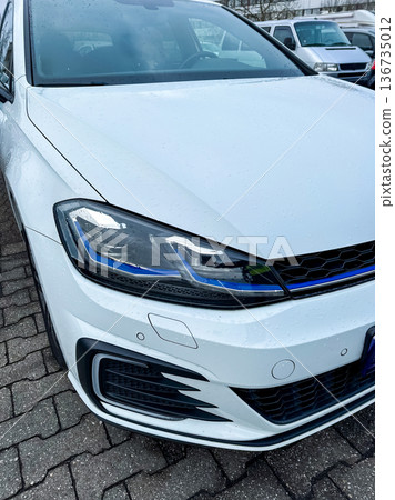 New car parked in a lot with wet pavement and cloudy weather showing details of the headlight and front grille while no other vehicles are nearby 136735012
