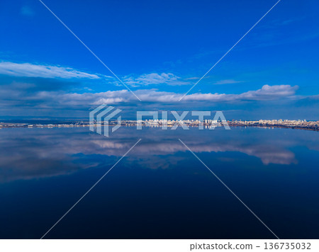 Calm water reflects blue sky and city skyline during midday at the lakeshore Calm water reflects blue sky and city skyline during midday at the lakeshore 136735032