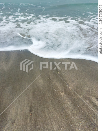 Waves wash over the sandy beach during late morning hours near the shoreline, creating patterns in the sand and foam as water retreats back into the ocean Waves wash over the sandy beach during late morning hours near the shoreline, creating patterns in the sand and foam as water retreats back into the ocean 136735043