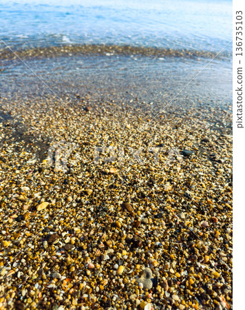Close-up view of small pebbles and sand along the shore of a beach during a sunny day with water gently washing over the shore Close-up view of small pebbles and sand along the shore of a beach during a sunny day with water gently washing over the shore 136735103