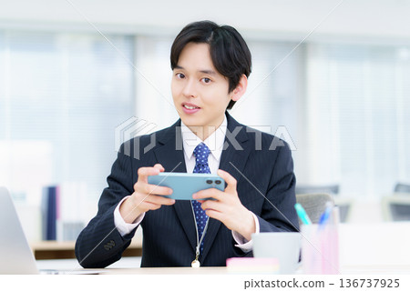 Smiling young Japanese man watching videos on his smartphone in the office Smiling young Japanese man watching videos on his smartphone in the office 136737925