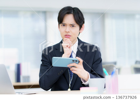 A young Japanese man with a troubled face watching videos on his smartphone in the office A young Japanese man with a troubled face watching videos on his smartphone in the office 136737926