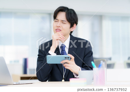 A young Japanese man with a troubled look on his face holding a smartphone in his office A young Japanese man with a troubled look on his face holding a smartphone in his office 136737932