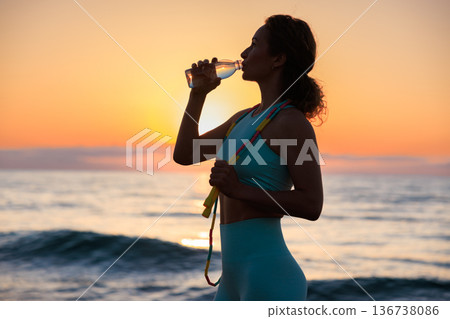 Fit woman drinking water at the beach after running outdoors in the morning. Sporty female relaxing after training. 136738086
