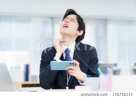 Young Japanese man scratching his neck while watching videos on his smartphone in the office 136738215