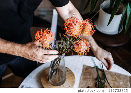 Person places orange pincushion protea into glass vase on table. sustainable weddings, eco-friendly events, biodegradable decor, low-impact floristry Person places orange pincushion protea into glass vase on table. sustainable weddings, eco-friendly events, biodegradable decor, low-impact floristry 136738825