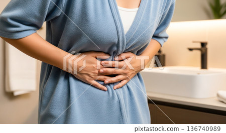 Woman in blue bathrobe holds her stomach, experiencing discomfort in a modern bathroom with sink and towels visible in the background 136740989