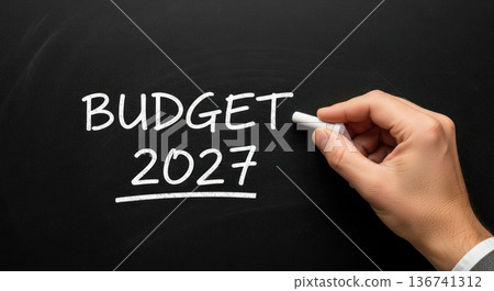 Hand writing "BUDGET 2027" on a black chalkboard with white chalk, emphasizing financial planning and future projections in a professional setting 136741312