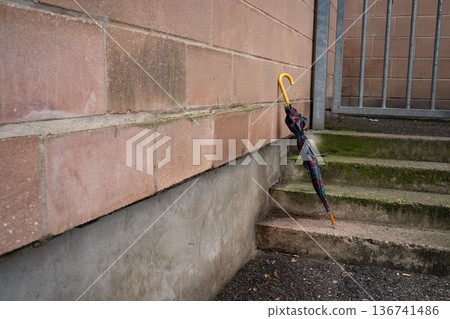 Plaid umbrella on stone stairs leaning against an industrial wall. Everyday urban scene showing a personal accessory, representing mobility, casual style, and outdoor life. Plaid umbrella on stone stairs leaning against an industrial wall. Everyday urban scene showing a personal accessory, representing mobility, casual style, and outdoor life. 136741486