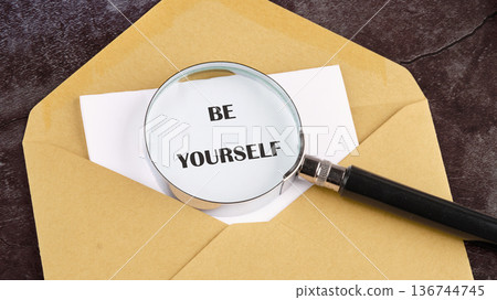 A gleaming magnifying glass rests on a white sheet, revealing the encouraging words BE YOURSELF, nestled within a golden envelope upon a textured dark surface 136744745
