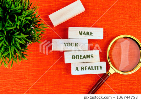 MAKE YOUR DREAMS A REALITY in wooden blocks on orange, with green foliage and a golden magnifying glass MAKE YOUR DREAMS A REALITY in wooden blocks on orange, with green foliage and a golden magnifying glass 136744758