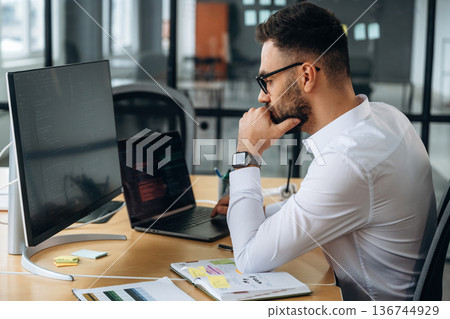 View from the side. Business person is sitting by computer in office and working View from the side. Business person is sitting by computer in office and working 136744929