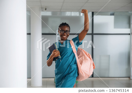 Arm is raised up, celebrating and smiling. Woman student with pink backpack is indoors 136745314
