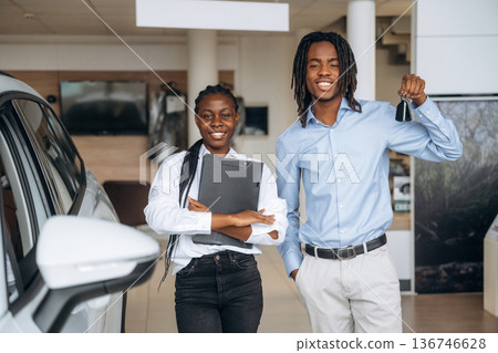 Happy owner is holding keys. Car seller is assisting client in the automobile salon 136746628