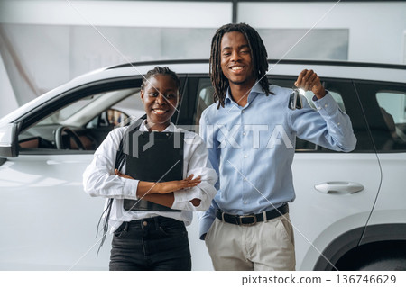 Happy owner is holding keys. Car seller is assisting client in the automobile salon Happy owner is holding keys. Car seller is assisting client in the automobile salon 136746629