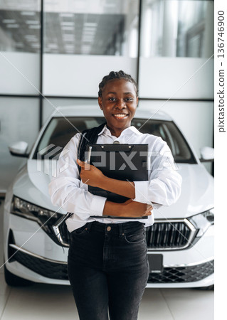 Black notepad in hands, smiling. Car seller, businesswoman is standing in automobile salon Black notepad in hands, smiling. Car seller, businesswoman is standing in automobile salon 136746900