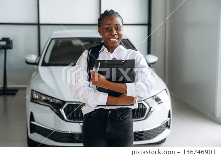 Black notepad in hands, smiling. Car seller, businesswoman is standing in automobile salon Black notepad in hands, smiling. Car seller, businesswoman is standing in automobile salon 136746901