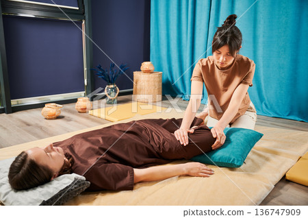 Masseuse making thai yoga massage. Woman therapist doing traditional massage treatment, stretching female patient's body. Client lying on mat, enjoying hand massage in calm atmosphere. 136747909