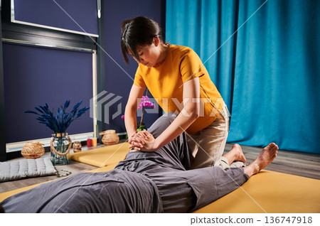 Masseuse making thai yoga massage. Woman therapist doing traditional massage treatment for male patient indoors. Client lying on mat, meditating, enjoying hand massage in calm atmosphere. 136747918