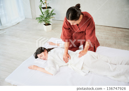 Masseuse making thai yoga massage. Woman therapist doing traditional massage treatment for female patient indoors. Client lying on mat, meditating, enjoying hand massage in calm atmosphere. 136747926