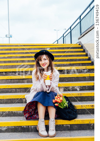 Portrait of young smiling woman with fresh tulips and coffee cup sitting on stairs at the city street enjoying the moment. Urban street fashion. Springtime yellow mood. Happiness and positive vibes Portrait of young smiling woman with fresh tulips and coffee cup sitting on stairs at the city street enjoying the moment. Urban street fashion. Springtime yellow mood. Happiness and positive vibes 136749419