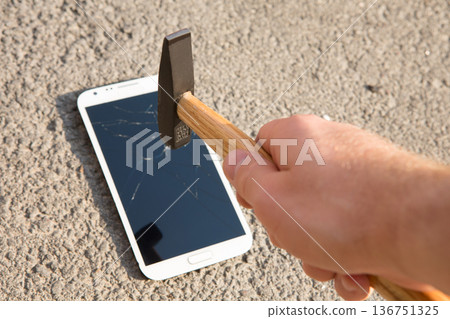 A hand drives a large metal nail through a smartphone screen with a hammer 136751325