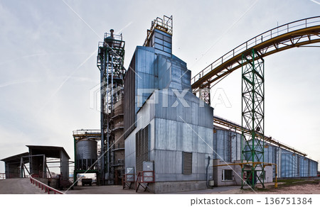 Industrial grain processing plant with steel silos and conveyor systems under a blue sky. Industrial grain processing plant with steel silos and conveyor systems under a blue sky. 136751384