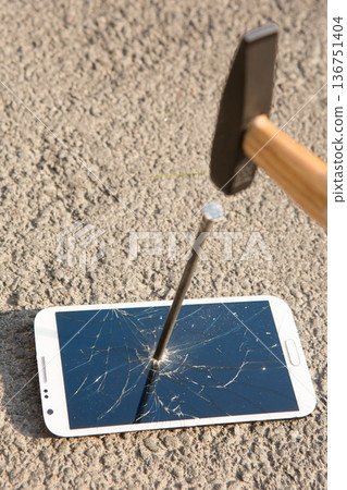 A hand drives a large metal nail through a smartphone screen with a hammer 136751404