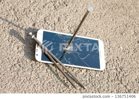 A white smartphone with a shattered screen pierced by a large metal nail on asphalt 136751406