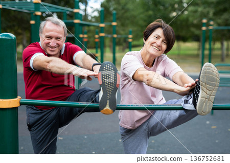 Mature couple doing leg stretching on crossbar together on sports ground of city park 136752681