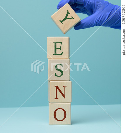 A hand in a blue medical glove places a wooden cube with the letter Y on a vertical stack of cubes, forming the word YES. 136752865