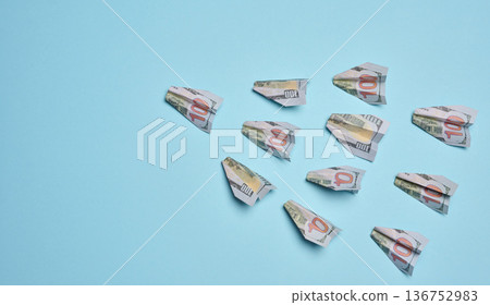 Paper airplanes made of hundred-dollar bills flying in one direction, against a blue background. 136752983