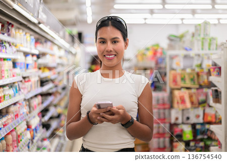 Smiling Woman Texting on Smartphone While Shopping in Supermarket Aisle 136754540