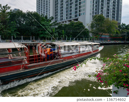 Passenger boat moving on canal water in city with green trees and flowers on bank, public water transportation system and commuting travel. Passenger boat moving on canal water in city with green trees and flowers on bank, public water transportation system and commuting travel. 136754602