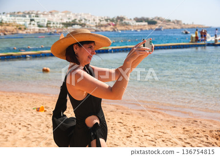 A female tourist in a hat films the beach at a hotel on her phone 136754815