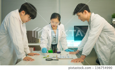Three science collaborate around a lab table, analyzing plant samples and chemical solutions together 136754990