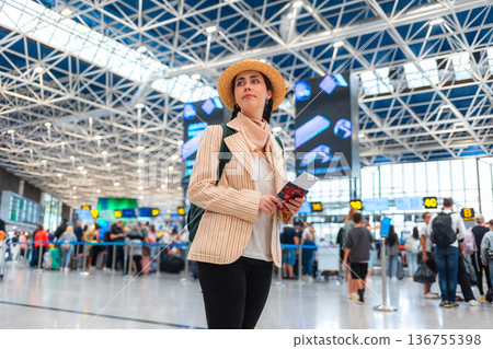 Bottom view of Caucasian young woman in straw hat smiling and holds passport. Defocused people in background. The concept of vacation and flight trip 136755398